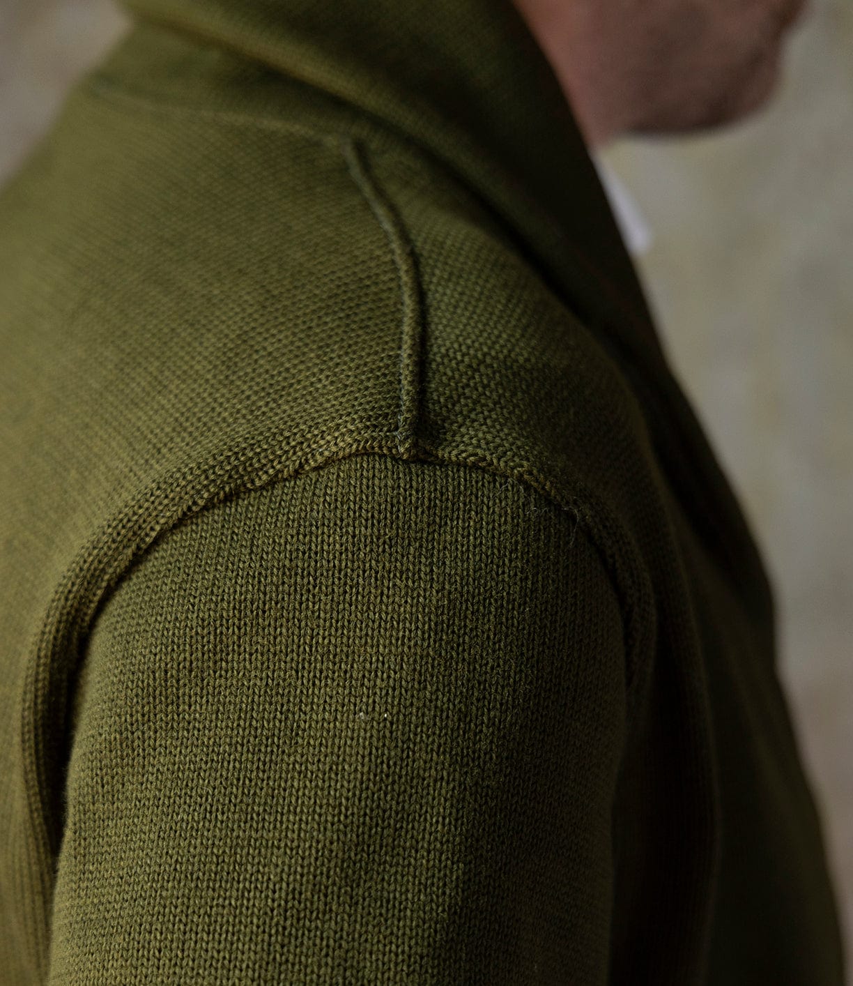 A close-up of a person's shoulder wearing the Best Made Co. Dehen Shawl Neck Sweater in moss green, featuring textured knit wool and a visible seam, with part of the neck and jawline shown.