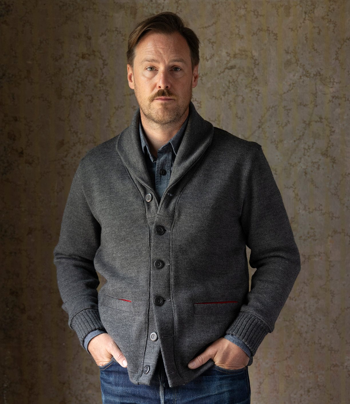 A man with light brown hair and a beard faces the camera, hands in pockets, wearing the Best Made Co. Dehen Shawl Neck Sweater in Charcoal over a denim shirt and blue jeans, set against a textured beige background inspired by classic American workwear.