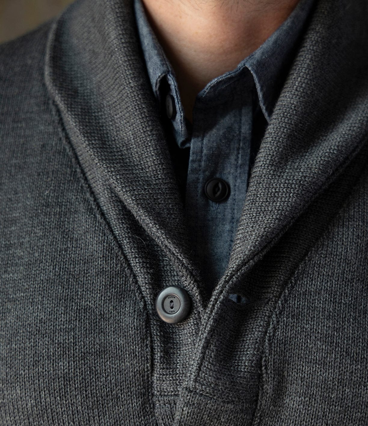 Close-up of a person wearing the Best Made Co. Dehen Shawl Neck Sweater in charcoal over a dark gray button-up shirt. The textured cardigan with large buttons recalls classic American workwear style; only the neck and upper torso are visible.