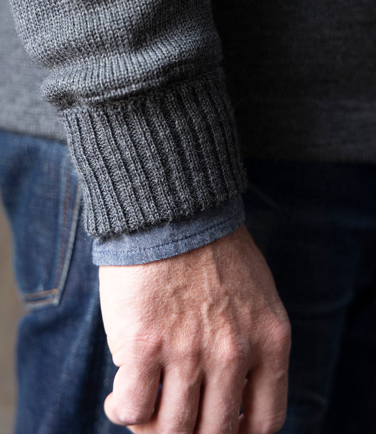 A close-up of a hand and wrist wearing the Best Made Co. Dehen Shawl Neck Sweater in Charcoal over a blue shirt sleeve, paired with blue jeans for a timeless American workwear look.