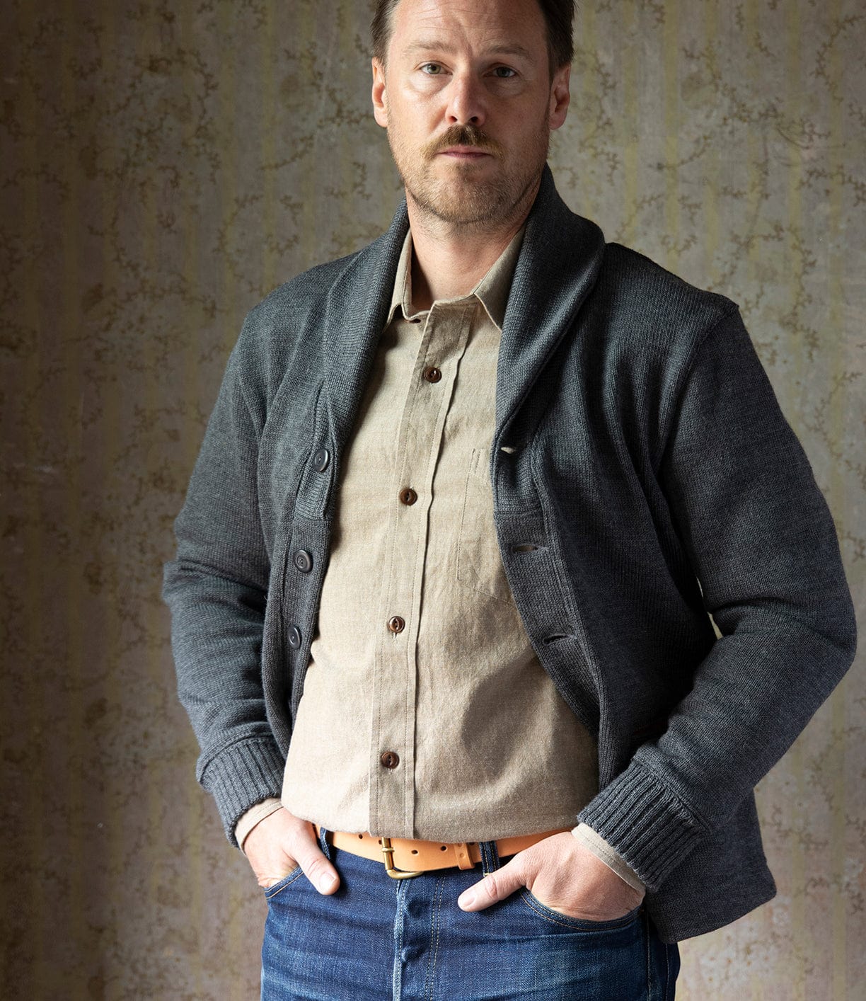 A man with light brown hair and a mustache stands indoors, hands in his pockets, wearing the Best Made Co. Dehen Shawl Neck Sweater in Charcoal over a beige shirt and blue jeans with a tan belt, looking calmly at the camera.