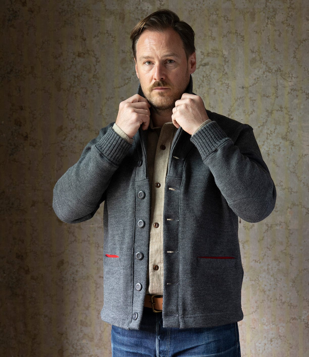 A man with light brown hair and a beard stands indoors, adjusting the collar of the Best Made Co. Dehen Shawl Neck Sweater in Charcoal over a beige shirt and jeans, looking directly at the camera against a patterned wall background.