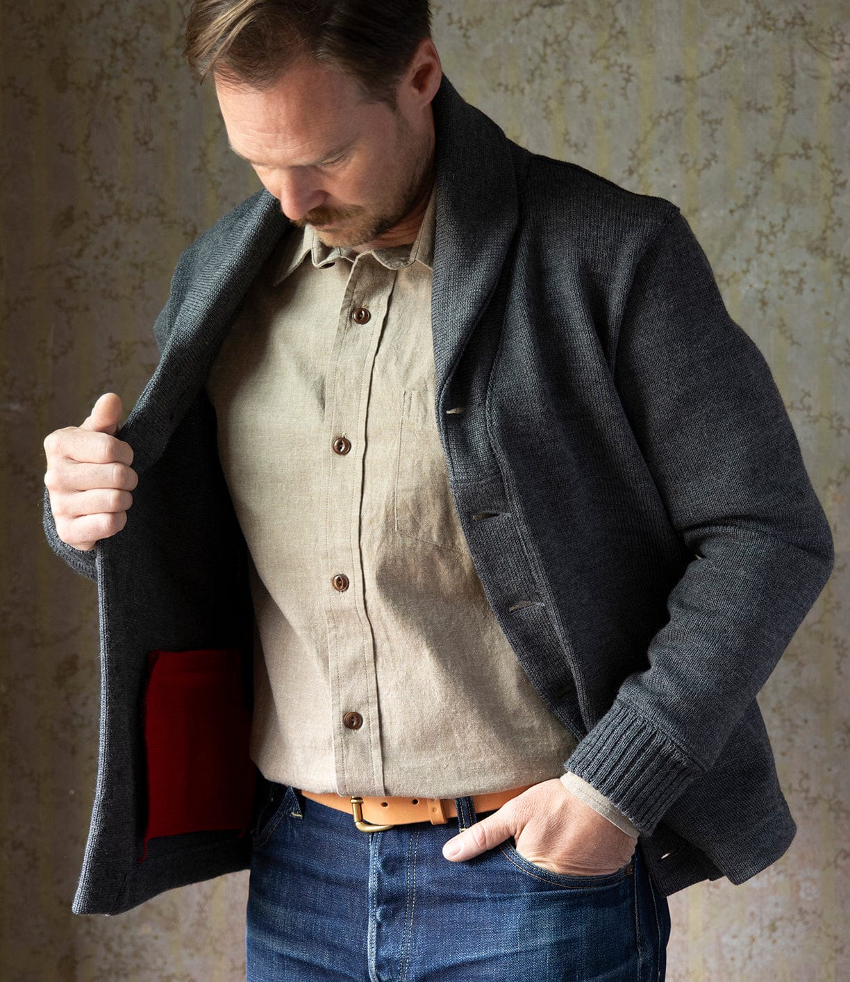 A man in a beige shirt and blue jeans opens the Dehen Shawl Neck Sweater Charcoal by Best Made Co., revealing a red inner pocket while looking down at it, set against a neutral patterned background.