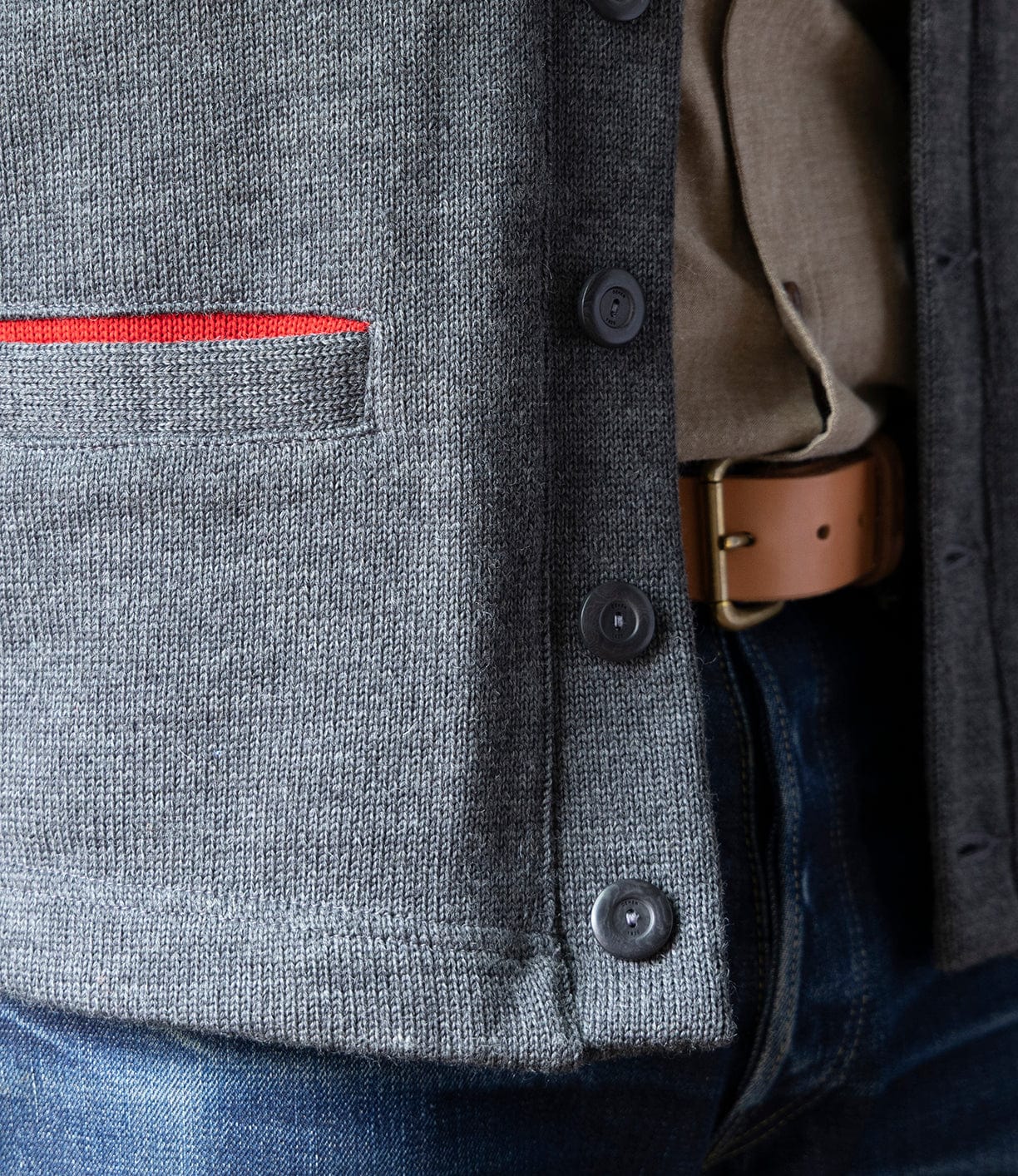 Close-up of a person in blue jeans, tan belt, beige shirt, and the Best Made Co. Dehen Shawl Neck Sweater Charcoal—a gray American workwear-inspired knit cardigan with buttons and a small red-accented pocket.