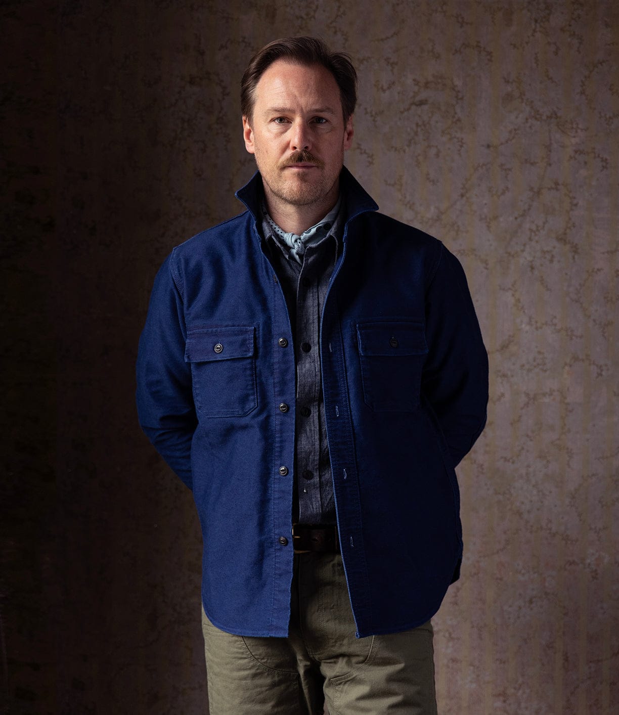 A man with light brown hair and a mustache stands against a textured beige background, wearing Best Made Co’s Indigo Moleskin Work Shirt under a blue jacket with olive green pants, his hands resting behind his back.