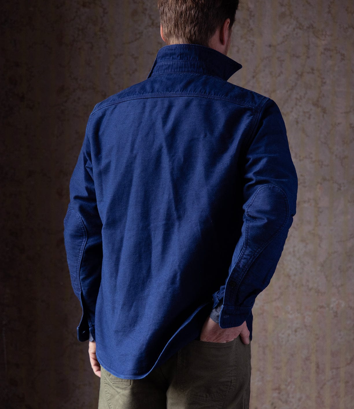A person faces away from the camera, wearing the Best Made Co Indigo Moleskin Work Shirt with collar turned up and olive green pants, set against a textured, neutral-toned background.