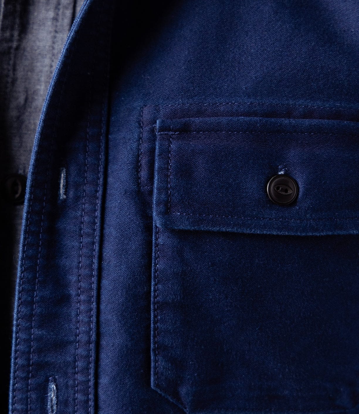 Close-up of the Best Made Co Indigo Moleskin Work Shirt, showing its high-density fabric, buttoned chest pocket, and triple-needle stitching. Layered over a gray shirt with a black button peeking out beneath.