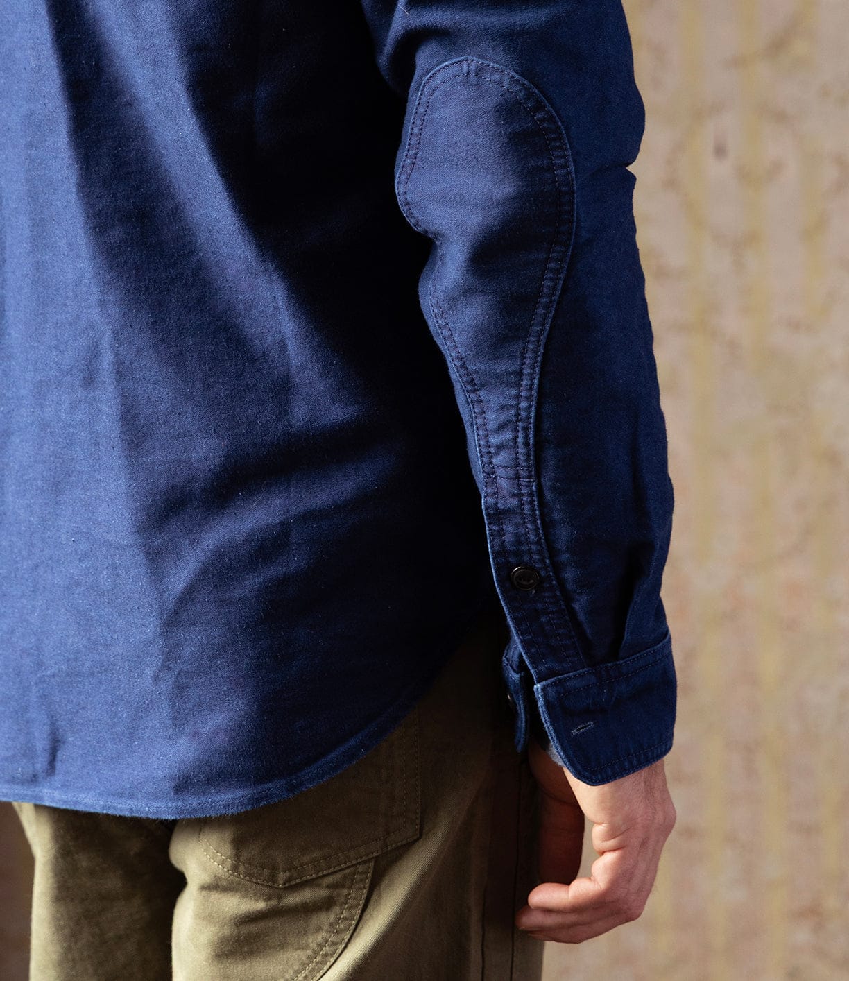 A close-up of a person’s arm wearing the Best Made Co Indigo Moleskin Work Shirt with visible stitching and buttoned cuff, paired with olive green pants. The high-density fabric pops against a softly textured beige background.