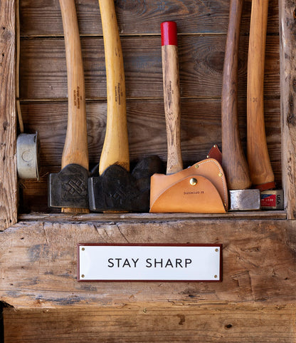Five axes and hammers with wooden handles hang on a rustic wall above the Best Made Co. “Stay Sharp” Enamel Steel Sign, highlighting the sign’s durable enamel and craftsmanship alongside rugged tools.