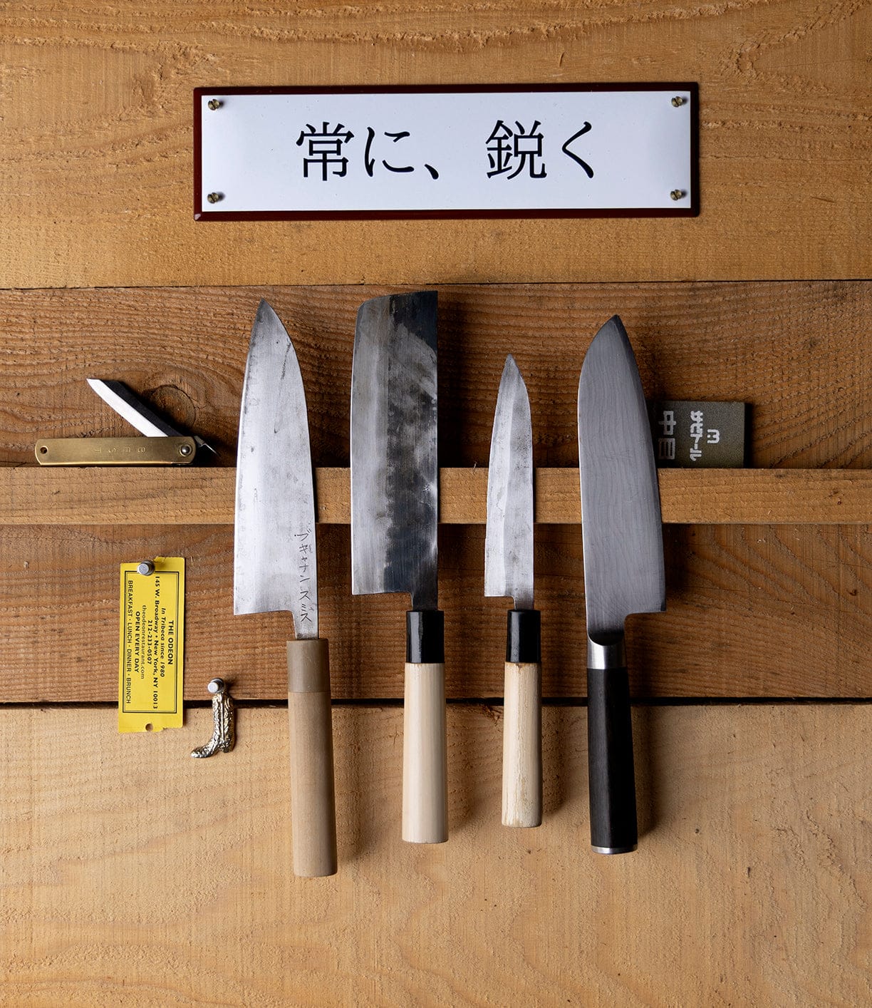 A wooden wall features five Japanese blades, a folding pocket knife, sharpener, and yellow tag below a magnetic strip. Above them hangs Best Made Company Co.’s “Stay Sharp” Enamel Steel Sign (Kanji Edition).