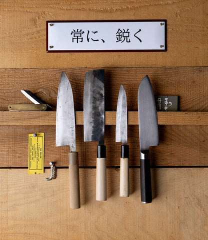 A magnetic knife rack on a wooden wall holds five Japanese blades, a small folding knife, a yellow sharpener, and the “Stay Sharp” Enamel Steel Sign (Kanji Edition) by Best Made Co. displayed above them.