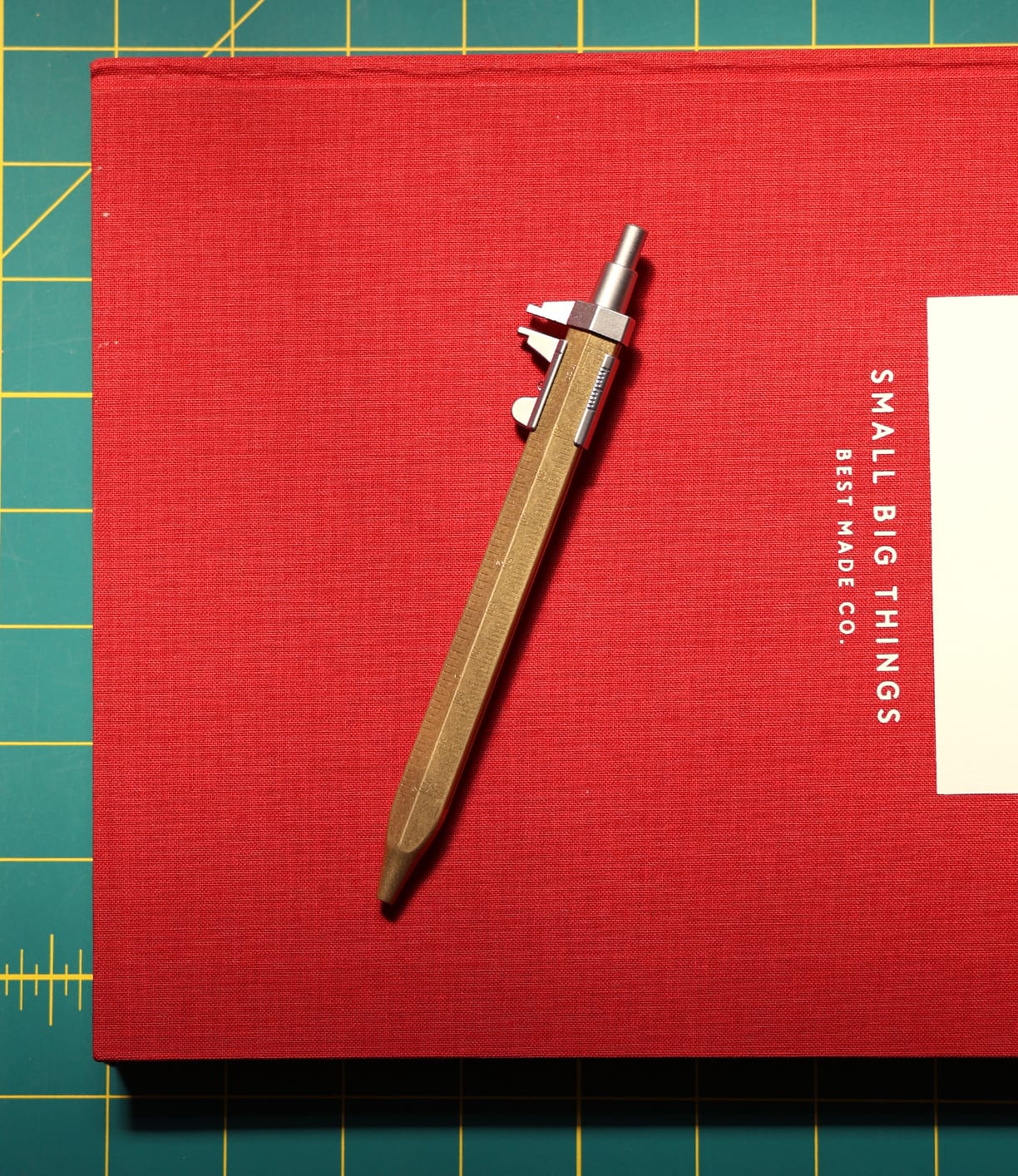 A Brass Messograf Pen by Best Made Co. rests diagonally on the red book titled SMALL BIG THINGS BEST MADE CO., which sits atop a green cutting mat with yellow grid lines.