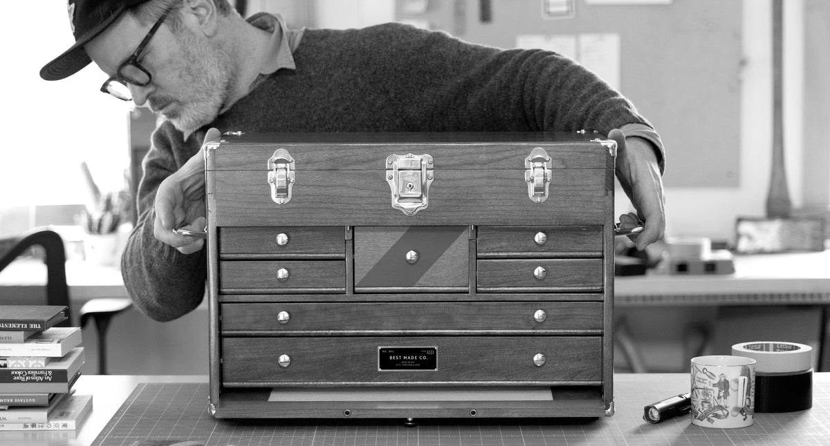 A person wearing glasses examines a wooden tool chest with multiple drawers on a desk. Various office items, books, and a cup are also visible in the workspace.