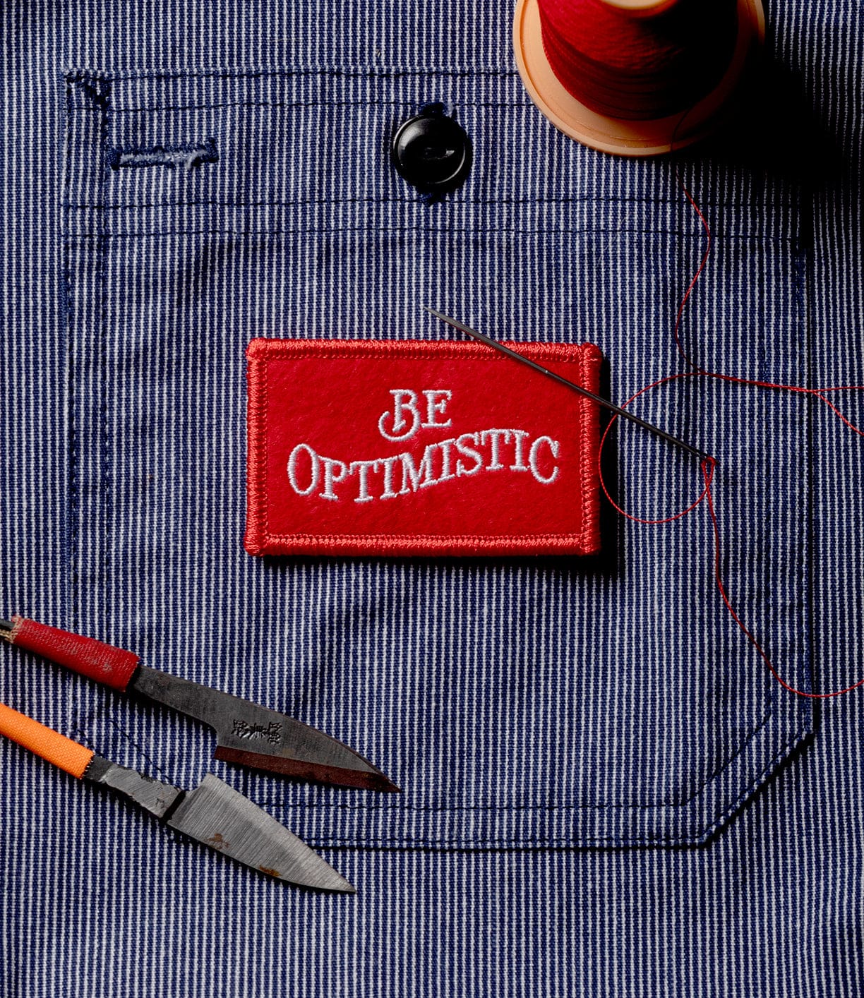 The Best Made Co Be Optimistic Patch, featuring "Be Optimistic" in white letters on red, sits with a needle, scissors, and red thread atop blue and white striped fabric—a practical addition to any survival kit.