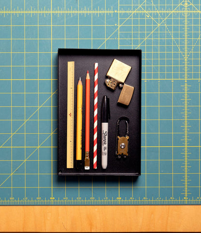 A Best Made Co. Spare Parts Tray (Matte Black) holds pencils, a ruler, a striped straw, a Sharpie, and padlocks on a blue and yellow cutting mat—ideal for organizing your workbench. Set includes three trays.