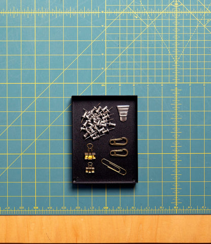 Best Made Co. Spare Parts Trays (Set of 3) in matte black hold silver push pins, binder clips, and safety pins on a green cutting mat with yellow grid—ideal for organizing your workbench.