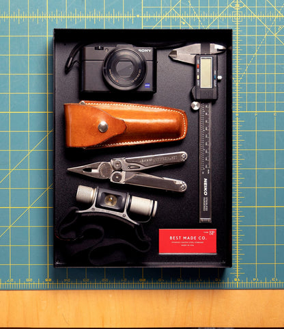 Best Made Co.'s Spare Parts Trays (Set of 3) in Matte Black keep essentials—like a digital camera, caliper, multitool, ruler, pouch, binoculars, and red Best Made Co. card—organized on your workbench or green cutting mat.