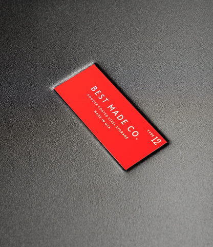 A red rectangular label on a textured dark surface reads, BEST MADE CO. Type 12 Strongbox - Matte Black. Made in USA in white capital letters, ensuring safe storage for your valuables.