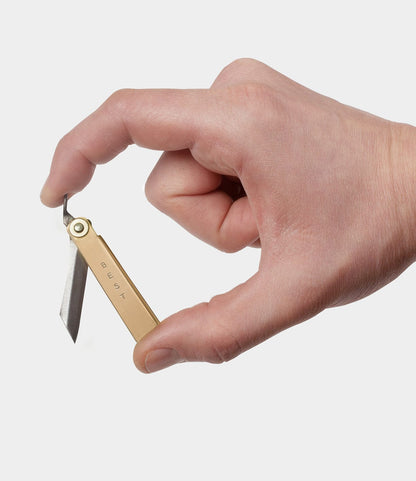 A hand holds the Mini Brass Higo Knife by Best Made Co. between thumb and index finger, with the blade partially opened and "BEST" engraved on its brass handle against a plain white background.