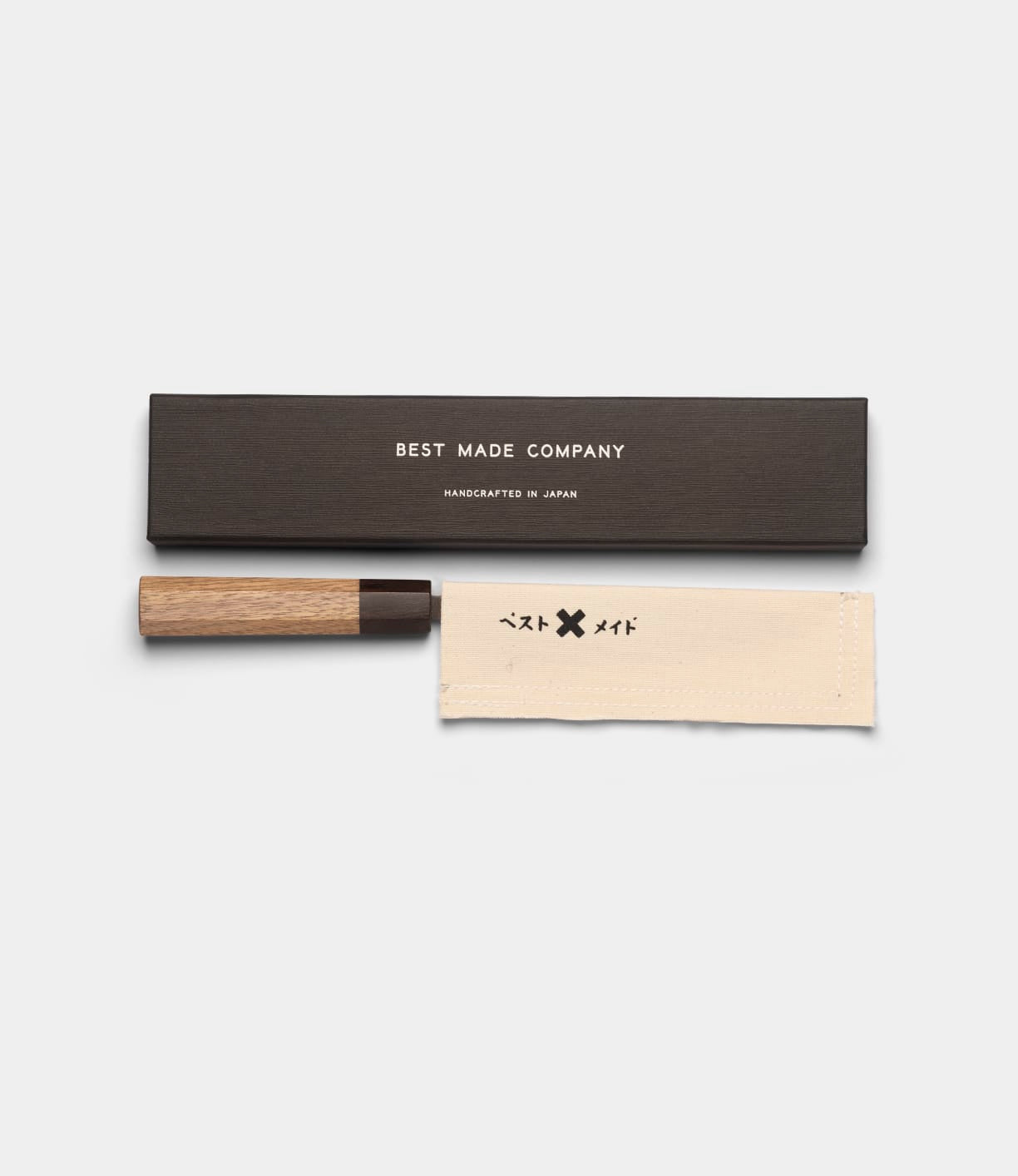 The Aogami Petty Knife by Best Made Co. features a wooden handle and a dark Blue Paper Steel No. 2 san-mai blade. It comes with a canvas sheath adorned with small black symbols for protection, and is presented in a black box labeled “Best Made Company, Handcrafted in Japan.”.