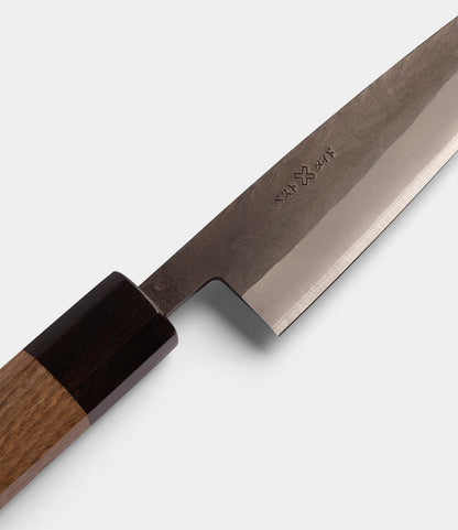 A close-up of the Best Made Co. Aogami Petty Knife, crafted with Blue Paper Steel No. 2 in a traditional san-mai construction, featuring a wooden handle and engraved characters on the blade, set against a plain light gray background.