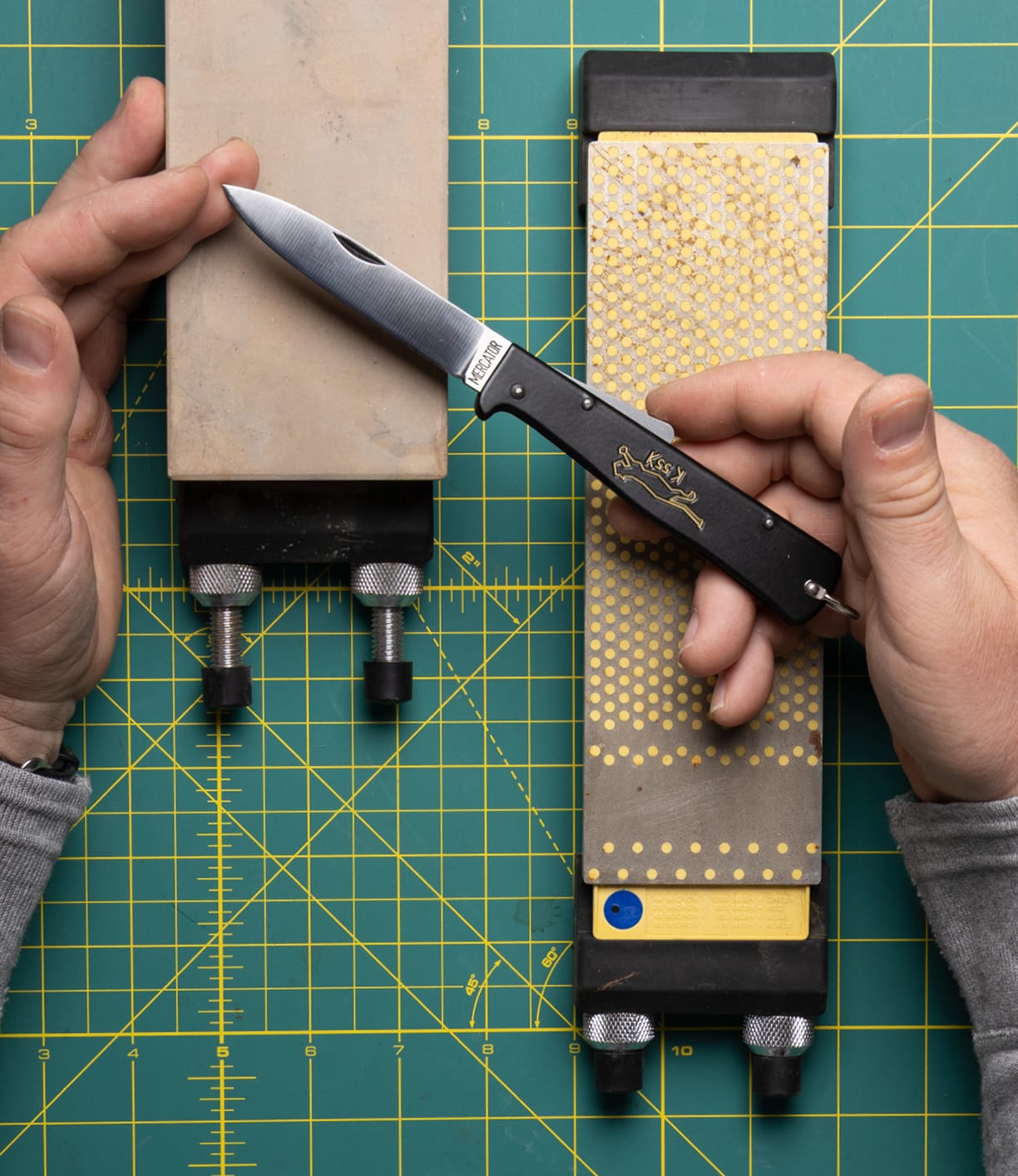 A person sharpens a Best Made Co. K55K Mercator carbon steel knife with a sharpening stone in one hand and another stone beside it, both on a green cutting mat with a yellow grid.