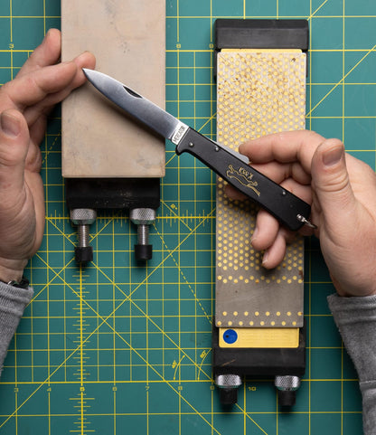 A person sharpens a Best Made Co. K55K Mercator carbon steel knife with a sharpening stone in one hand and another stone beside it, both on a green cutting mat with a yellow grid.