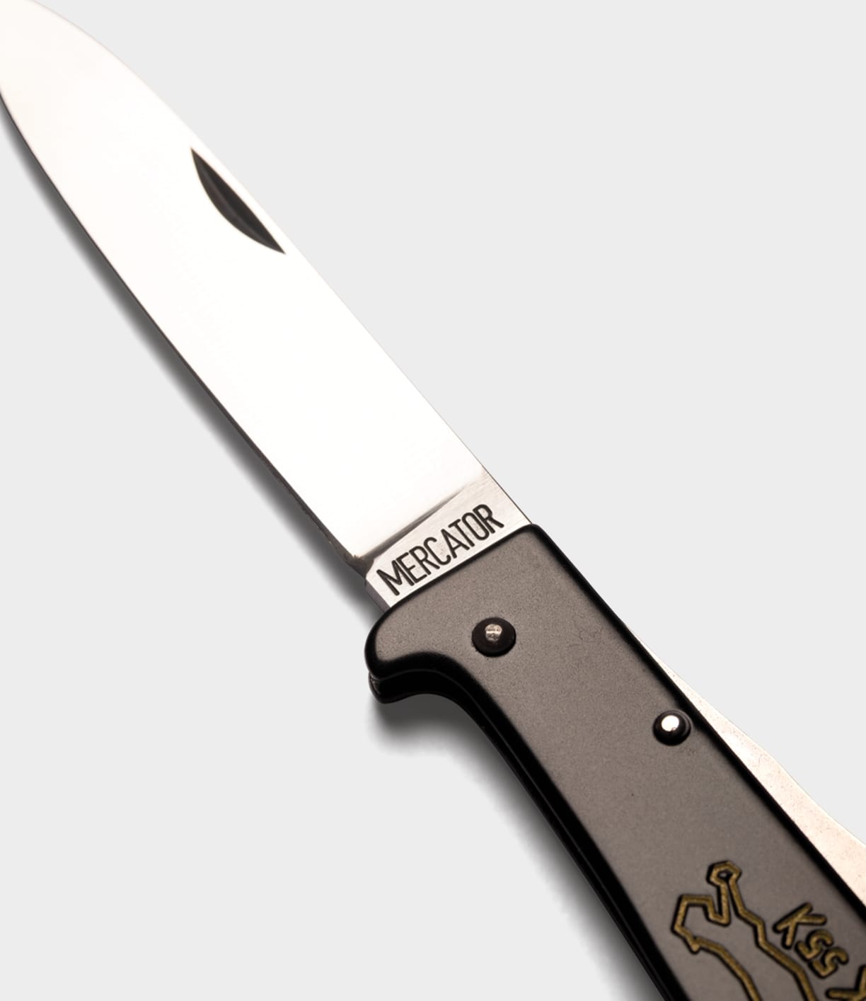 A close-up of an open K55K Mercator by Best Made Co., showing a silver blade engraved MERCATOR and a dark handle with a brass-colored cat design, photographed on a light gray background.