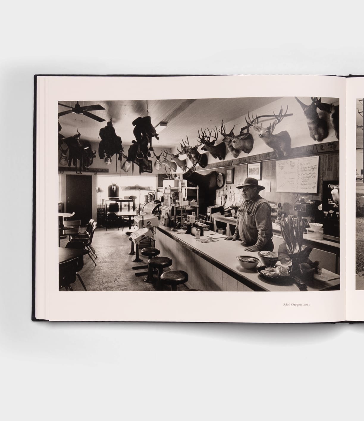 Black and white photo of a man behind a diner counter, walls with mounted animal heads. Empty stools face the counter; tables fill the space. "Open Range" by Best Made Co. evokes rustic charm, like a Robert Adams photography book.