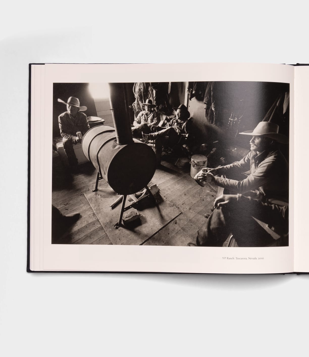 A black-and-white photo evokes the spirit of Open Range by Best Made Co., featuring four cowboys around a barrel stove in a rustic room filled with gear, capturing a warm, relaxed, and communal Western atmosphere.