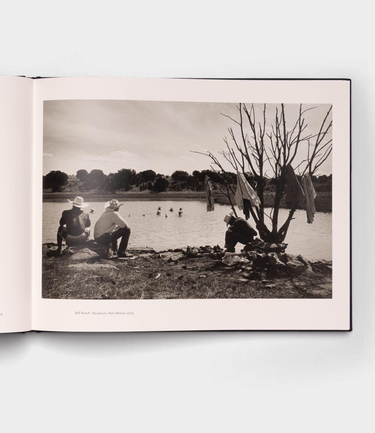 The Best Made Co. "Open Range" photography book lies open, showing a black-and-white image of three people—two with cowboy hats—by a lake, clothes on a bare tree, wooden boats nearby, and swimmers in the background.