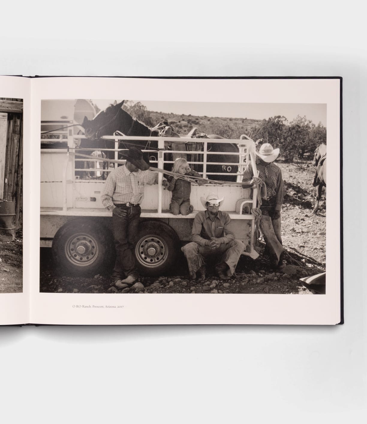 Open Range by Best Made Co.: A black-and-white photo inspired by Robert Adams, showing three adults and a child in cowboy hats and western attire beside a livestock trailer, with horses and open land in the background.