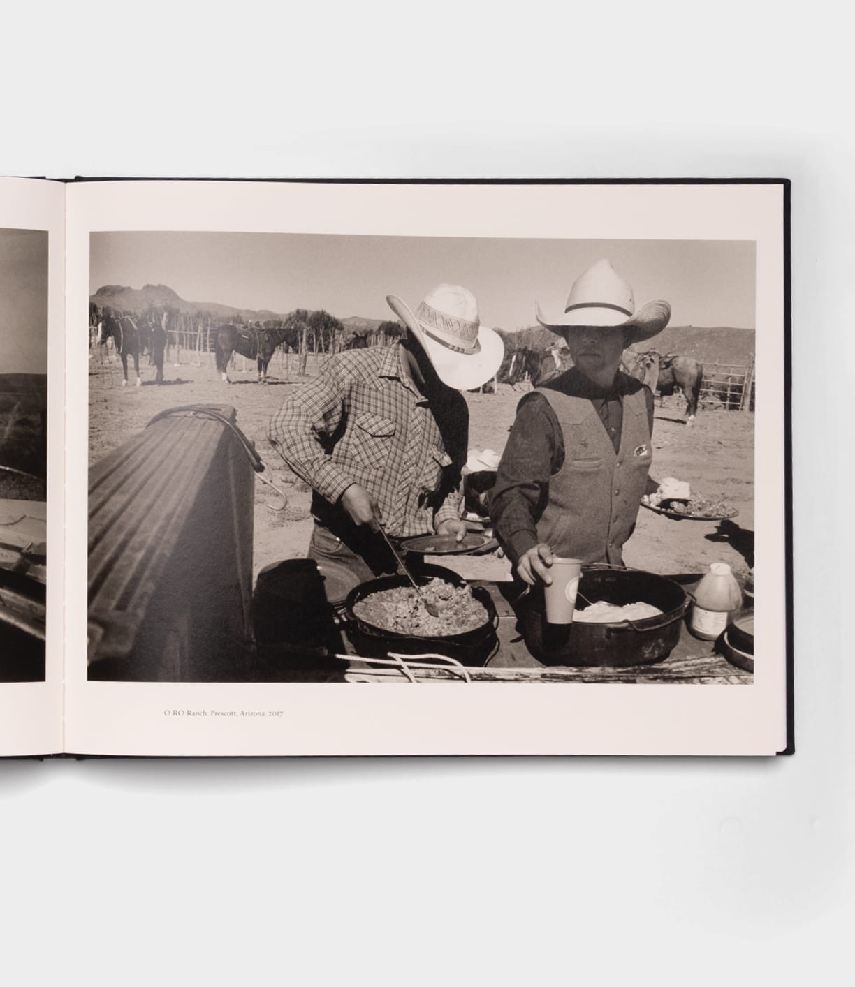 Black-and-white photo in the style of Robert Adams shows two cowboys cooking outdoors. One stirs a pan while the other pours a drink, with horses by a fence. Featured: "Open Range" by Best Made Co.
