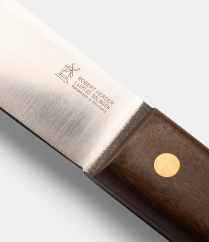 A close-up of the Best Made Co. Sailor’s Knife featuring a Brazilian Teak handle with a brass rivet. The Niolox steel blade is engraved with a windmill logo and the text: ROBERT HERDER, 1872 SOLINGEN, Handmade in Germany.
