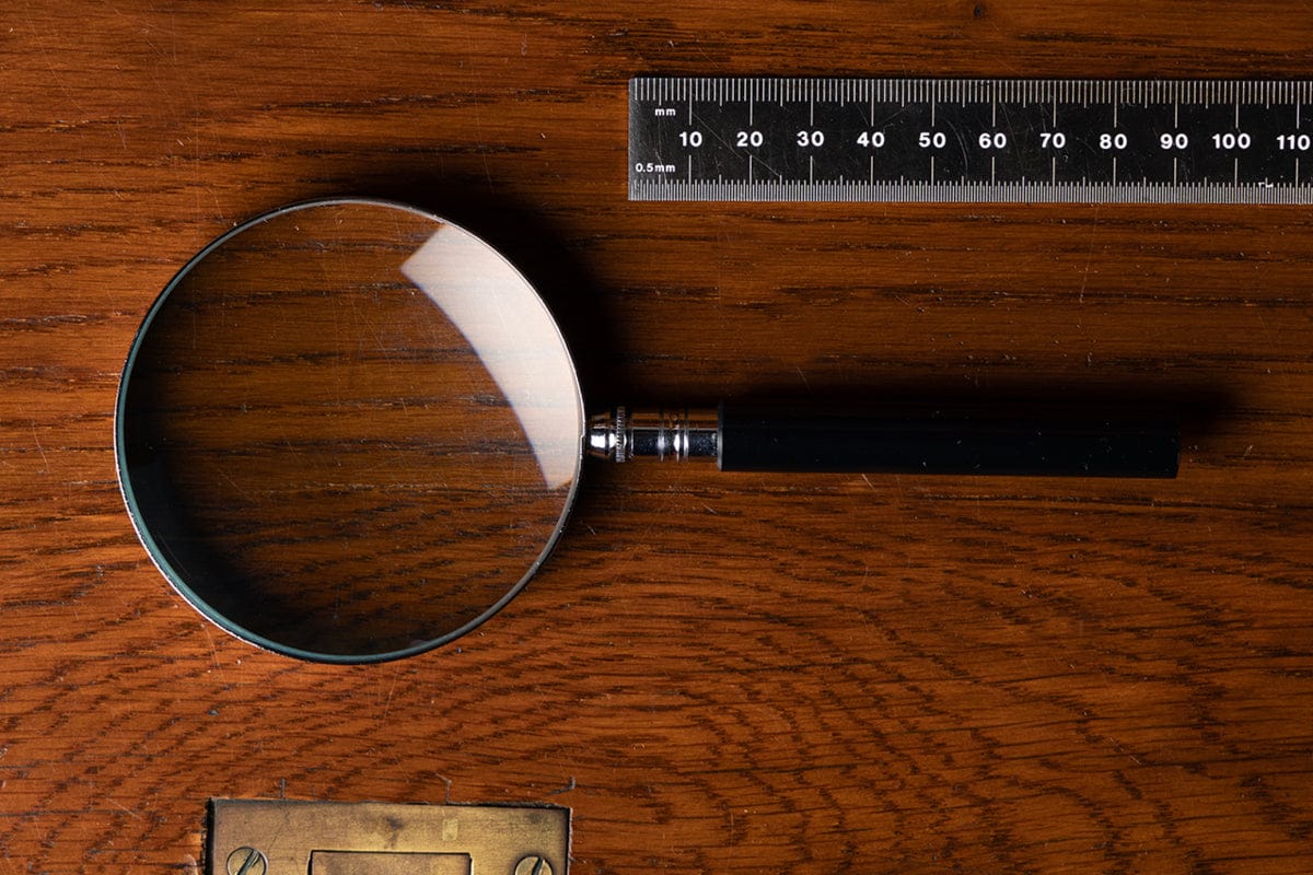 A magnifying glass and a black ruler with white markings are placed on a wooden surface. The magnifying glass is positioned to the left of the ruler.