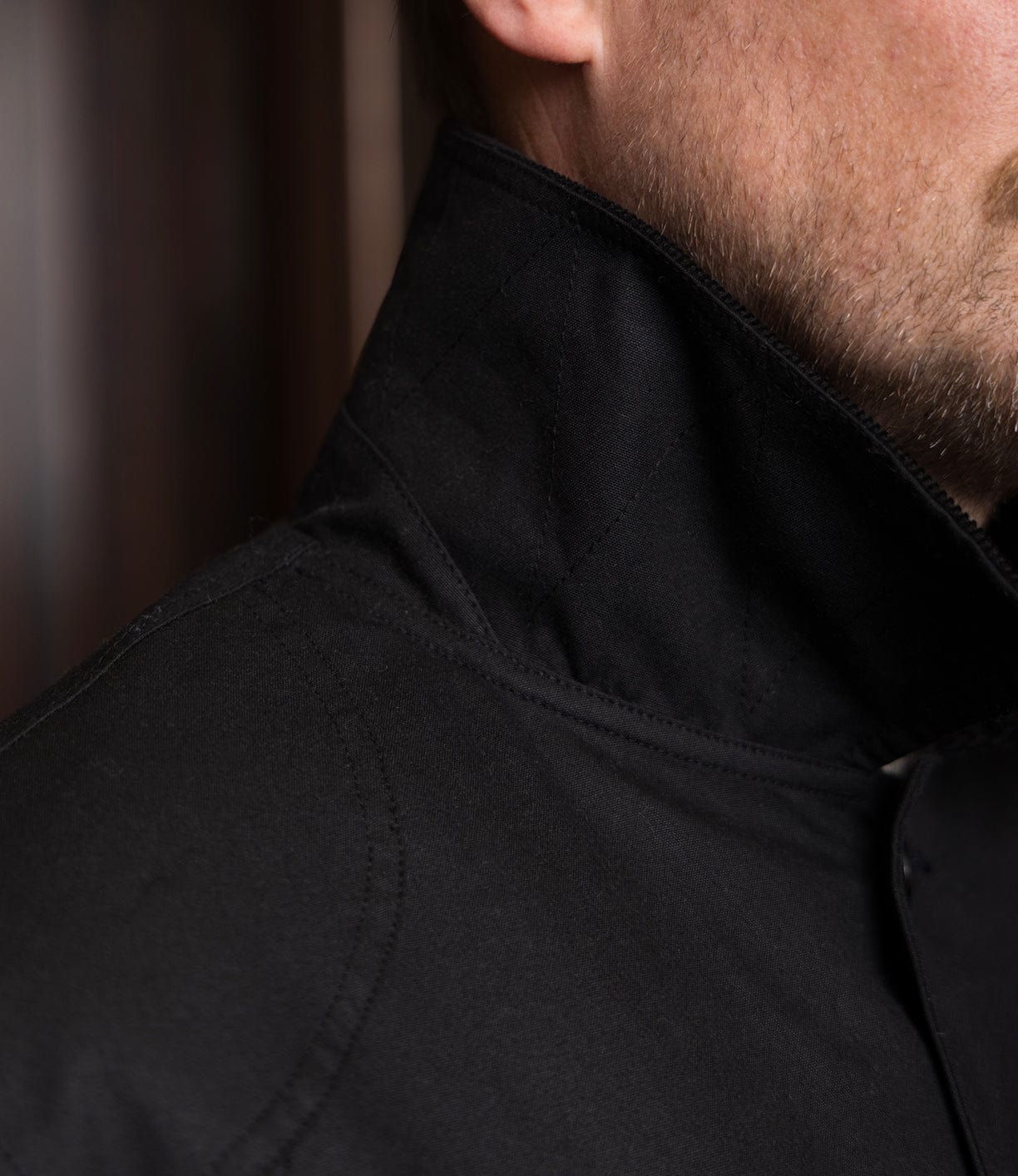 Close-up of a man's lower face and neck wearing the Best Made Co. Wool-Lined Waxed Field Jacket in black with the collar turned up, highlighting the weather-resistant fabric and detailed stitching.