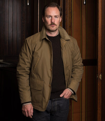 A man with light brown hair and a mustache stands indoors against wood paneling, wearing the Best Made Co. Wool-Lined Waxed Field Jacket in Tan over a dark sweater and jeans, one hand in his pocket.