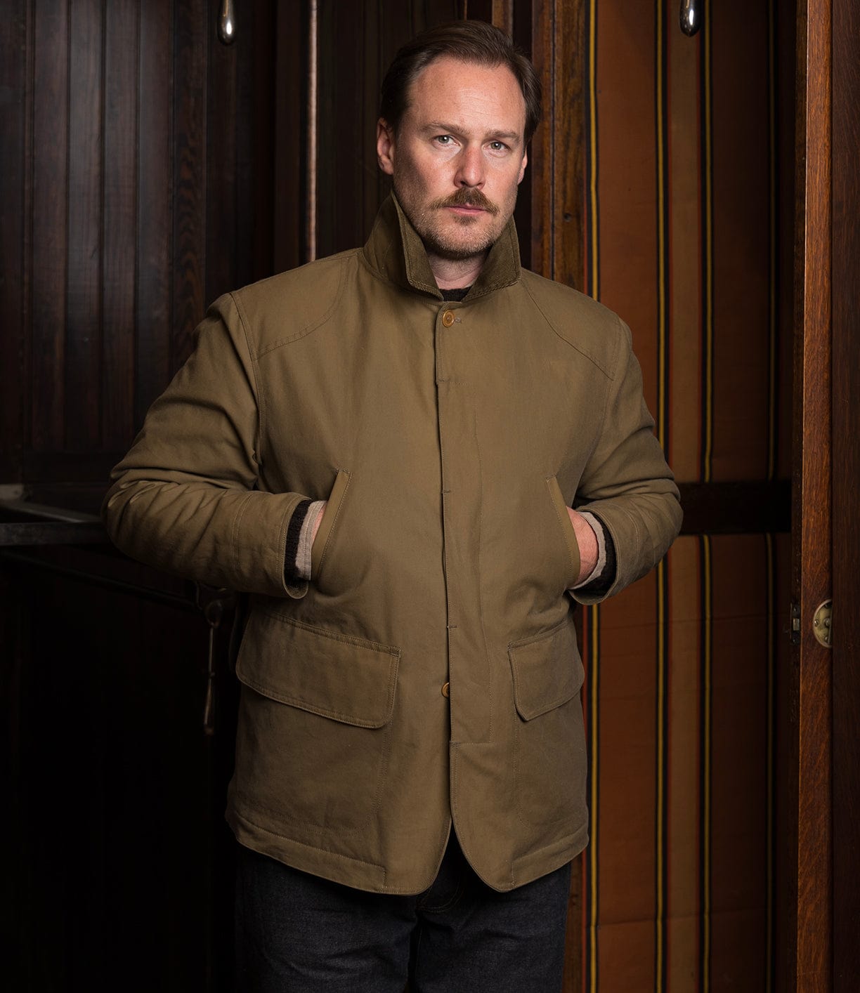 A man with light brown hair and a mustache stands indoors, hands in pockets, wearing the Best Made Co. Wool-Lined Waxed Field Jacket in Tan. Behind him are dark wooden walls and a striped curtain as he looks at the camera.