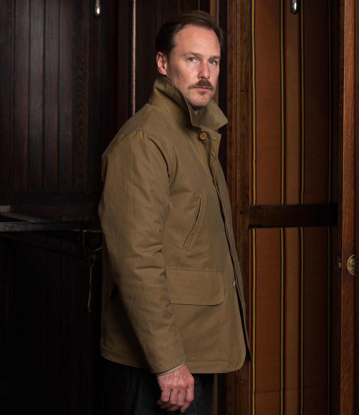 A man with light brown hair and a mustache stands indoors by dark wood panels and a striped curtain, wearing the Best Made Co. Wool-Lined Waxed Field Jacket in Tan, looking seriously at the camera.