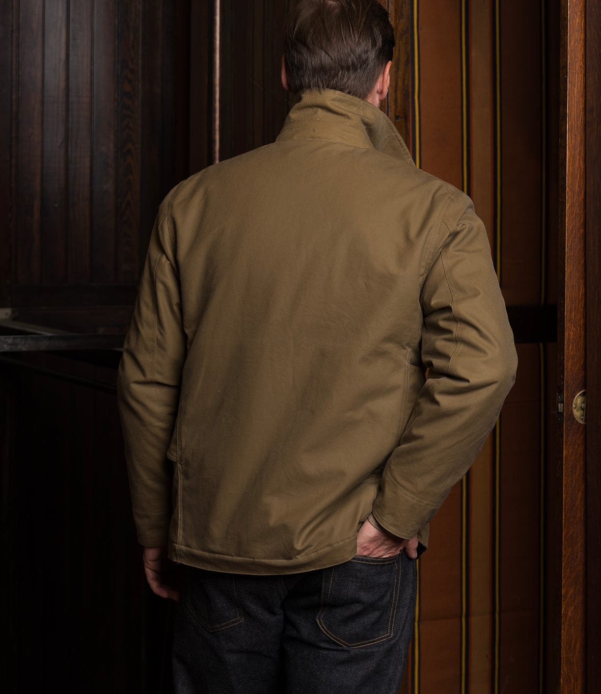A man stands indoors with his back to the camera, wearing the Best Made Co. Wool-Lined Waxed Field Jacket in Tan and dark jeans. The setting features dark wooden walls and a partially open door with vertical striped panels.
