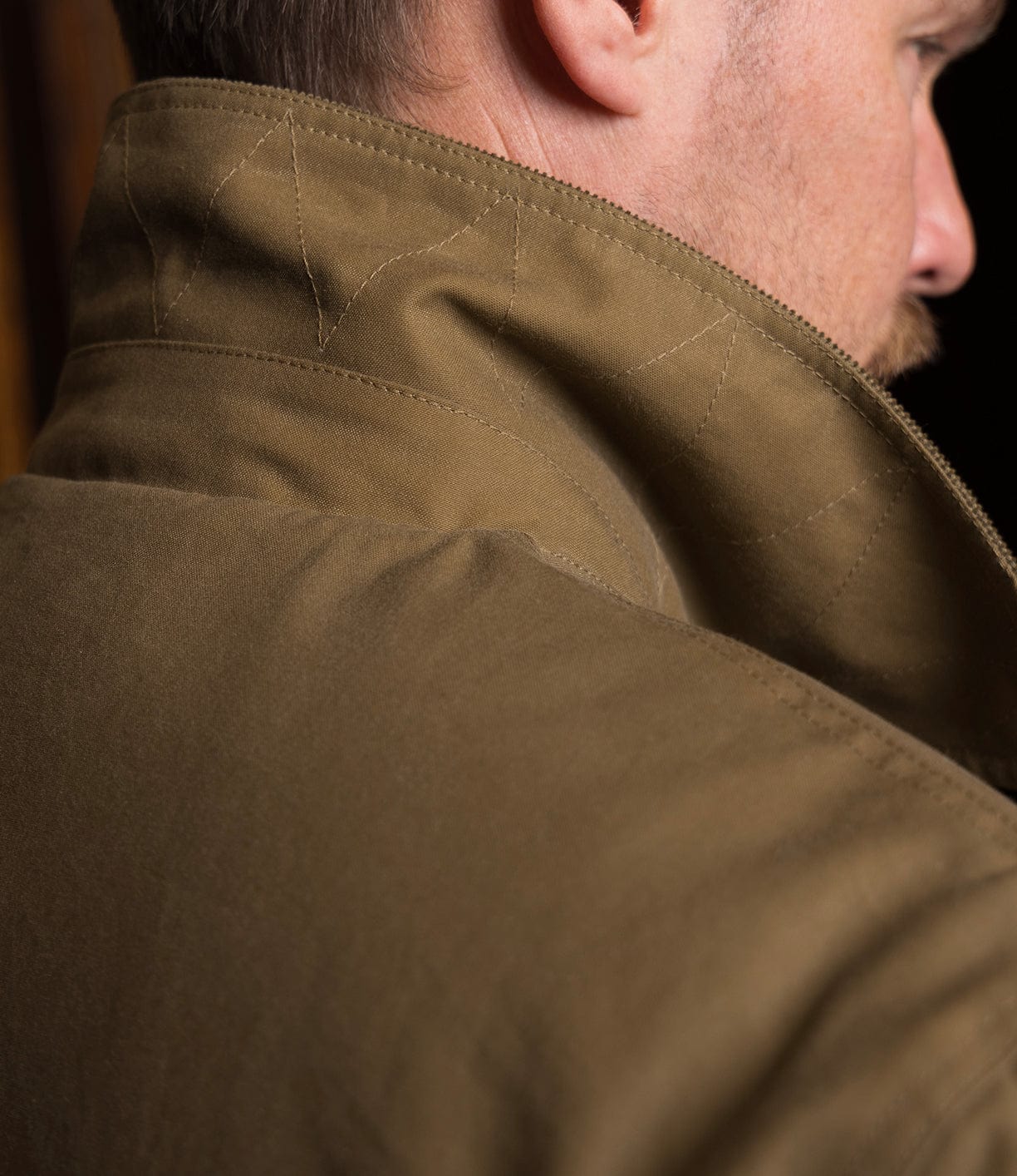 Close-up of a person in the Best Made Co. Wool-Lined Waxed Field Jacket Tan with the collar turned up, showcasing detailed stitching; face partially visible from the side.