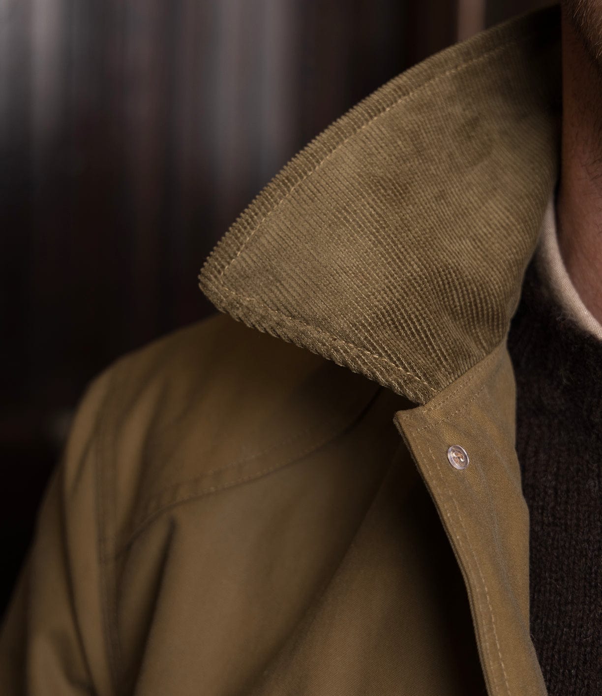 Close-up of a person wearing the Best Made Co. Wool-Lined Waxed Field Jacket in tan with the collar turned up, revealing its corduroy texture. A brown sweater is layered underneath; the background is dark and out of focus.