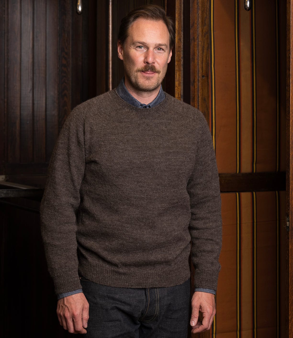 A man with light brown hair and a mustache stands indoors, wearing the Best Made Co. 5-Gauge Shetland Sweater over a blue collared shirt and dark jeans, with wooden panels and orange-striped walls behind him.