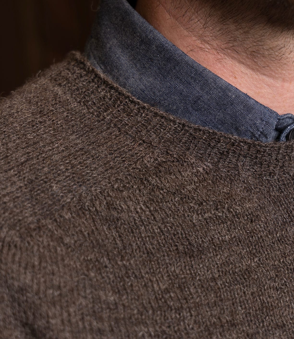 Close-up of a person's neck and shoulder in a blue collared shirt under the Best Made Co. 5-Gauge Shetland Sweater. This brown, textured knit provides lightweight warmth; the face is not visible.