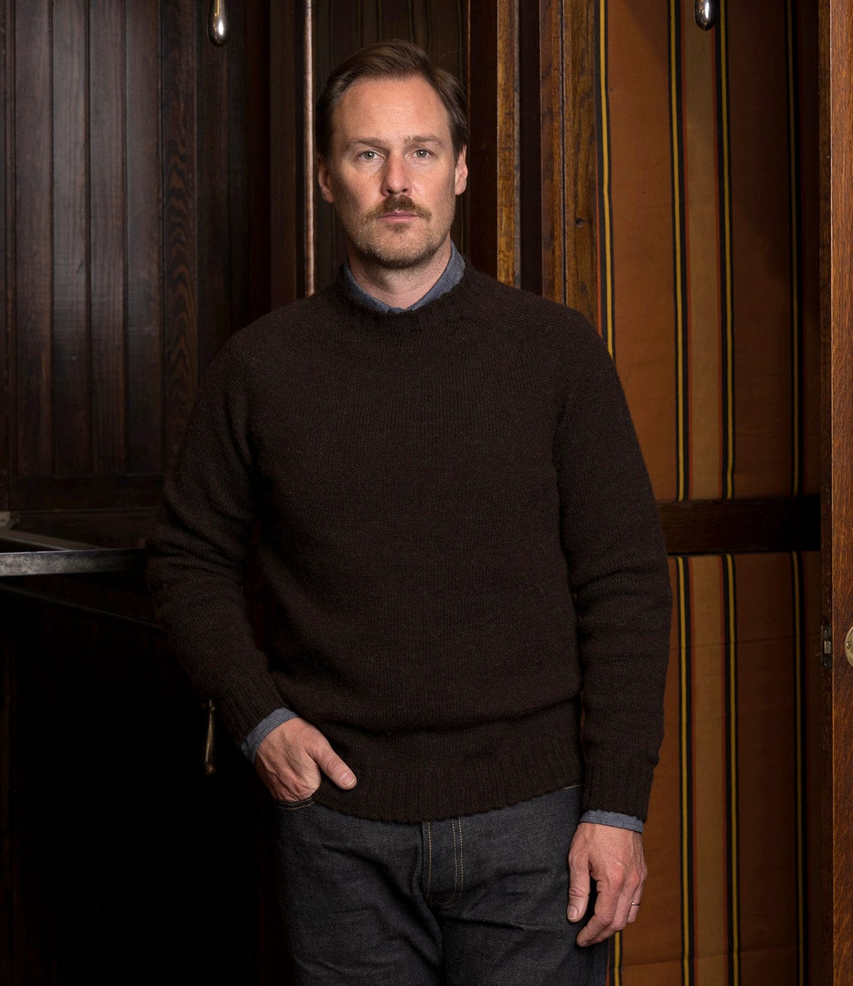A man with short hair and a mustache stands indoors, wearing the Best Made Co. Black Sheep Sweater over a collared shirt and dark jeans, one hand in his pocket, positioned before dark wood panels and striped curtains.