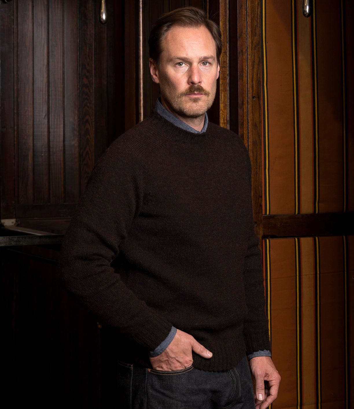 A man with light brown hair and a mustache, wearing a Best Made Co. Black Sheep Sweater over a collared shirt, stands indoors against dark wood paneling and striped walls, looking seriously at the camera.