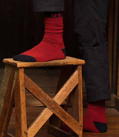 A person in dark pants models Best Made Co. Standard Wool Socks—red merino wool with black toes, heels, and cuffs—standing with one foot on a rustic wooden stool and the other on the floor for year-round comfort.