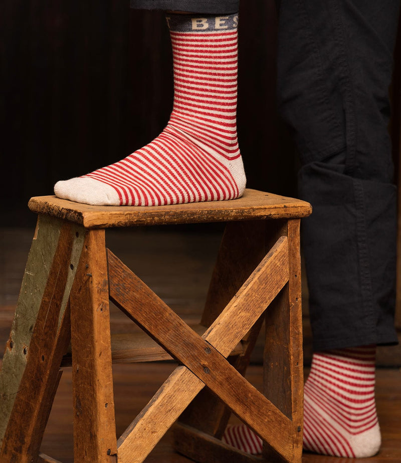 A person wearing red and white striped socks with the word BEST at the top stands on a wooden step stool. The person is dressed in dark pants and the background is dark and blurred.