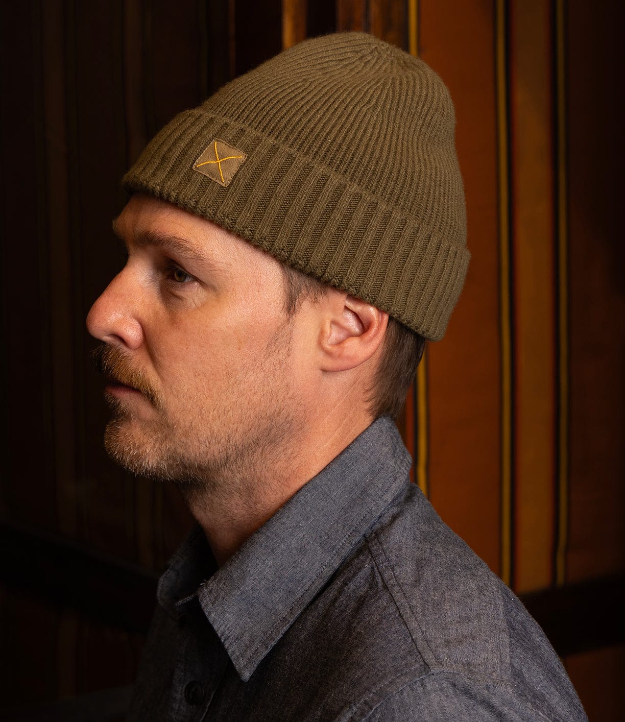 A man with light facial hair wears a ribbed olive green Cap of Courage by Best Made Co and a gray button-up shirt, looking left in a warmly lit room with striped brown and yellow walls.