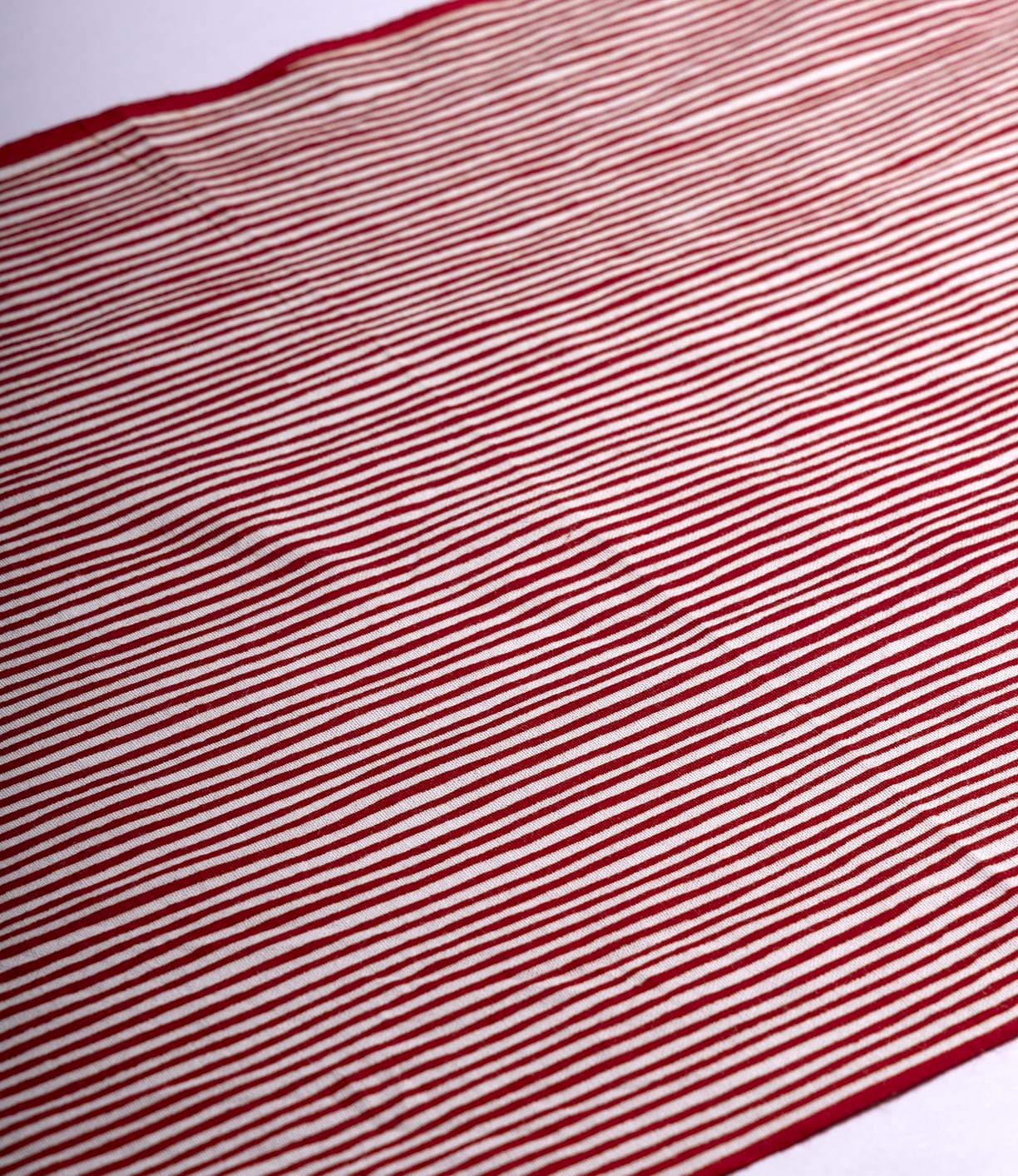 The Yoroke-Jima Tenugui by Best Made Co. features red and white horizontal stripes on hand-dyed Japanese fabric, laid flat with gentle wrinkles and shadows for a wabi-sabi feel. Evenly spaced stripes span the entire cloth.